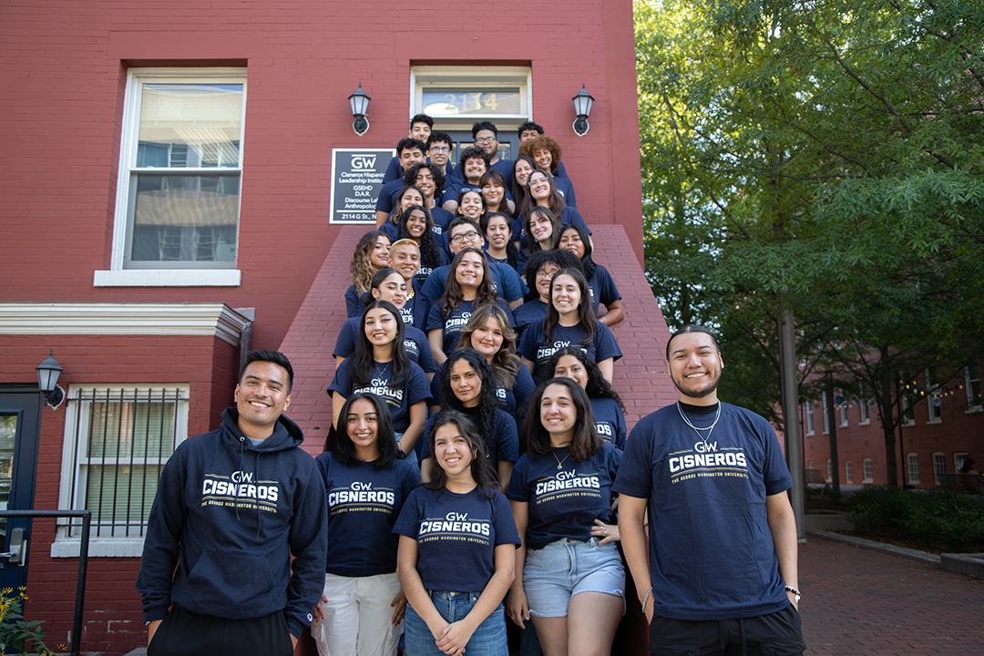 A group of students pose together on a staircase outside. They are all wearing blue shirts with writing