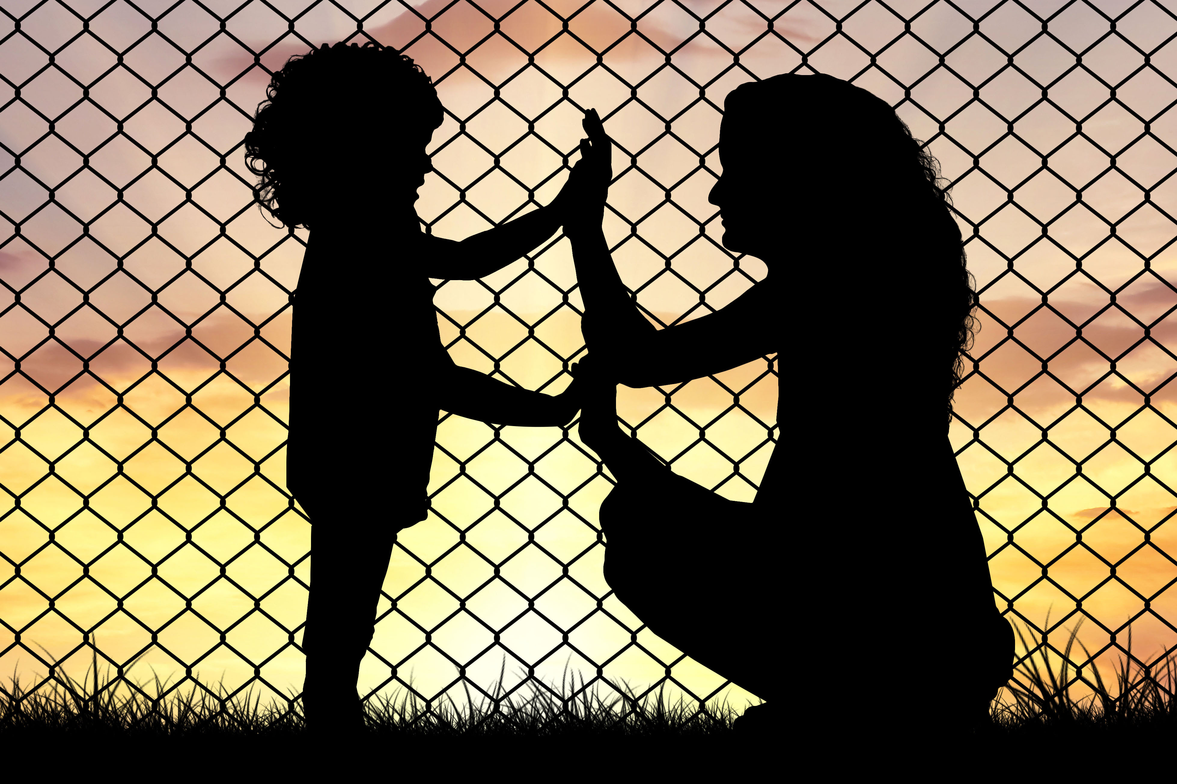 A silhouette of a mother and child in front of a fence holding hands