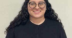 Cristy is a woman with round black glasses, a black button down sweater, and long dark, curly hair.