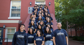 A group of students pose together on a staircase outside. They are all wearing blue shirts with writing